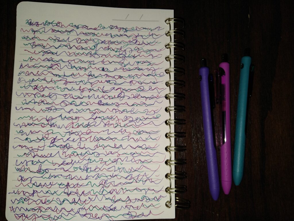 A lined notebook is open on a table next to three colored pens. The pens are purple, magenta, and teal. The notebook page is filled with overlapping squiggles of all three colors of the pens. 