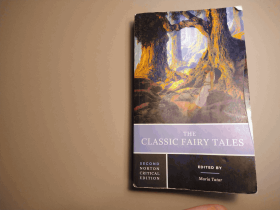 A hand opens the Norton book "The Classic Fairy Tales" edited by Maria Tatar. The opened book reveals a night sky turning to dawn with a moon and clouds inside it. 