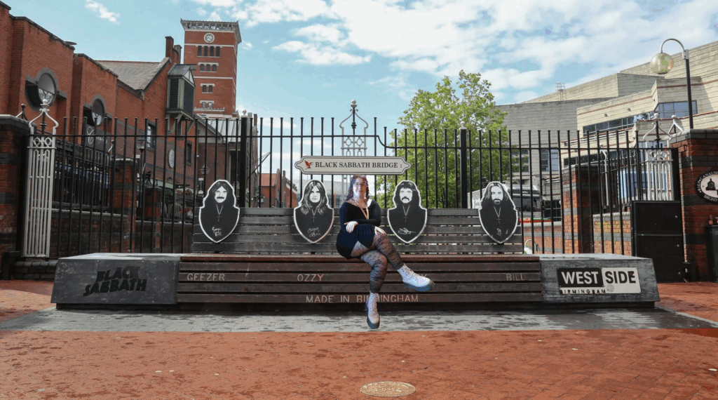 A biologically female person is edited to be sitting on the Black Sabbath Bridge Tribute Bench. They have pale skin, glasses, and purple hair. They are wearing all black except for their purple Doc Martin's boots.